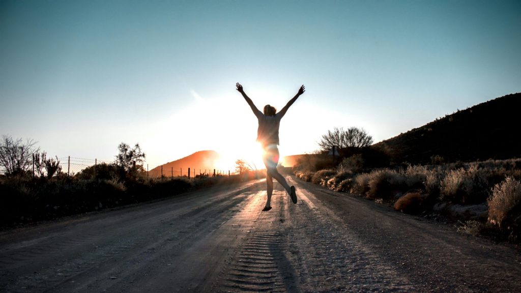 A joyful individual jumping on a dirt road during sunrise in a scenic desert landscape.